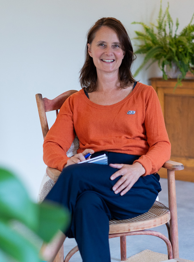 Portret van Isabel Janssens, glimlachend in een houten stoel met een notitieblokje en pen